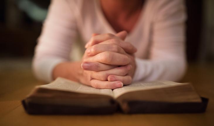 mulher sentada e apoiando as maos em forma de oração sobre uma biblia aberta