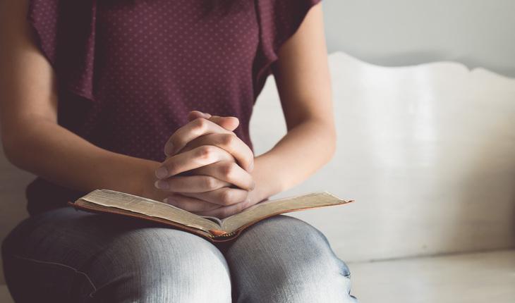 mulher sentada em um sofa segurando uma biblia no colo e com as maos em posicao de oraca
