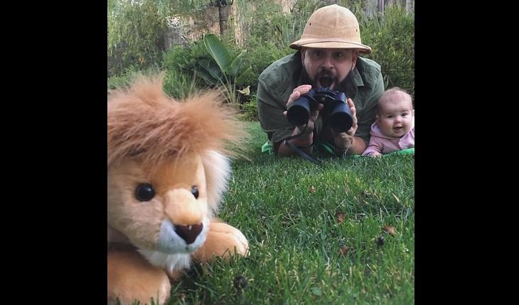 pai e filha fazendo uma fotografia engraçada, com ele de binoculo assustado e a filha ao lado rindo, no primeiro plano uma pelúcia de leão