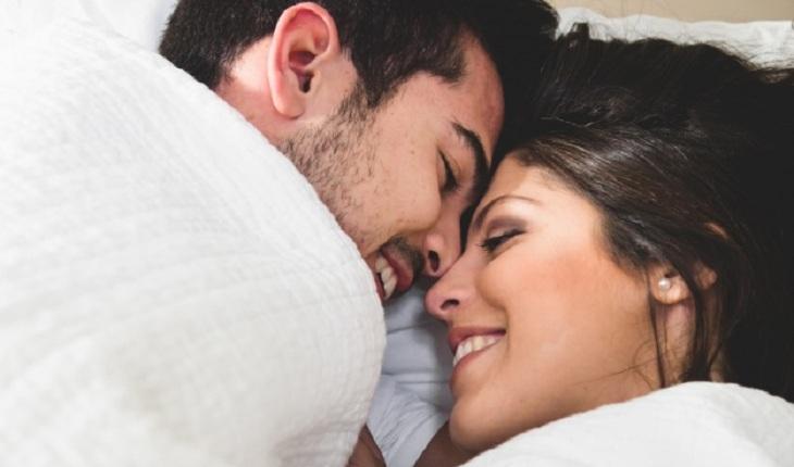 Na imagem, o casal está coberto com cobertor branco, com os olhos fechados e rosto encostando no outro sorrindo. Benefícios do sexo.