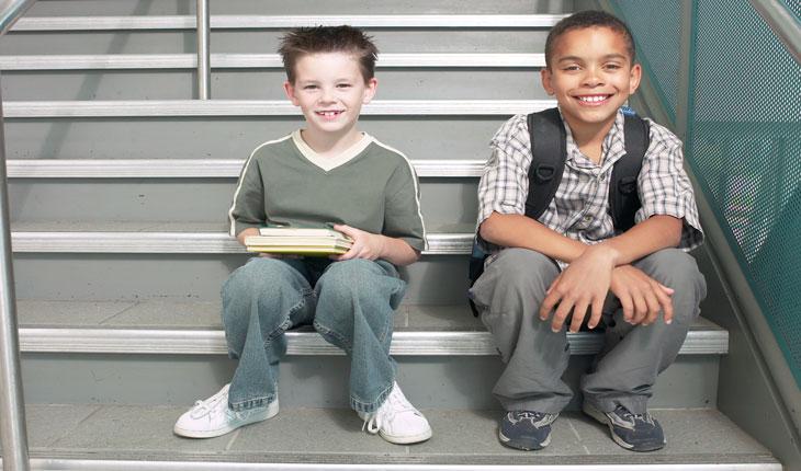 Na foto há dois meninos sentados em uma escada e segurando livros e cadernos.
