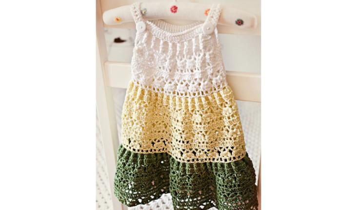 Na foto há um vestido de alça para criança feito de crochê. Ele é branco na parte do tronco, amarelo na parte da cintura e verde no barrado.