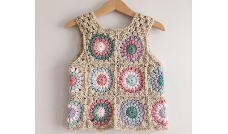 Na foto há um colete de crochê para bebês feito na cor bege, rosa-claro, azul-claro e outras cores suaves.