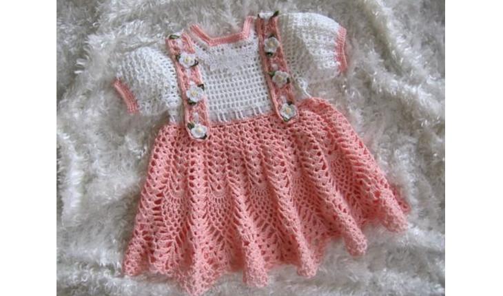 Na foto há um vestidinho de bebê feito de crochê. Ele tem as cores rosa e branco e têm detalhes de florszinhas.