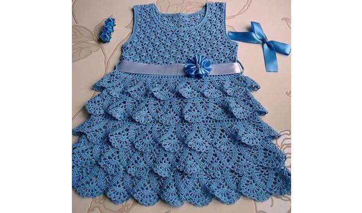 Na foto há um vestido de bebê feito de crochê na cor azul. A parte da saia é feita com babados. Na cintura há uma fita decorando.