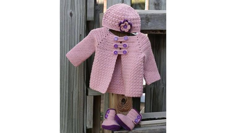 Na foto há um casaco rosa com detalhes em roxo feito de crochê. Há também uma bota rosa-claro e roxo e uma touca também rosa com um flor roxo.