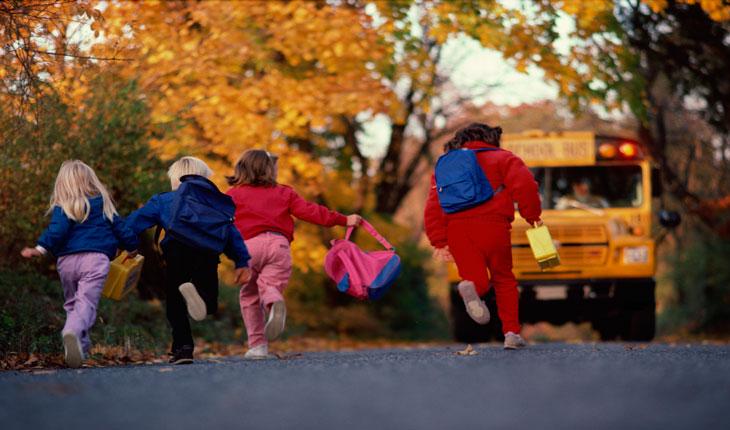 Na foto há várias crianças de costas correndo na rua com materiais de escola e mochilas na mão.