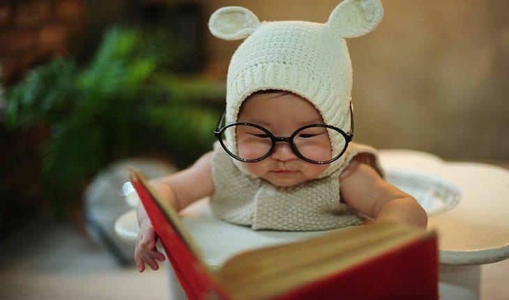 Imagem de um garoto com fenótipo asiático, usando uma roupa com touca com orelha de bichinho, óculos e lendo um livro.