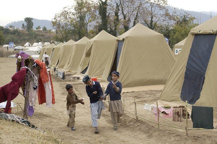 Na imagem, um acampamento de refugiados está montada com tendas em bege e três crianças andam e conversam na estrada de terra. Acolher os refugiados. 