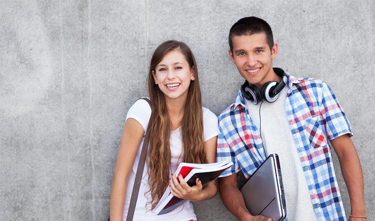 Na foto há um casal de adolescentes sorrindo para a foto e segurando livros e computadores.
