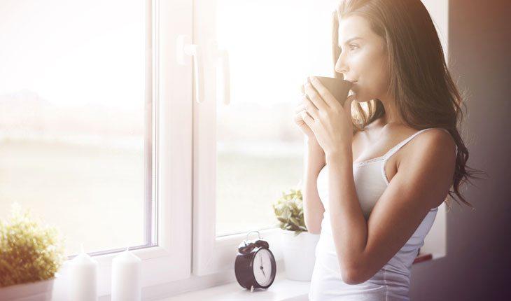 Na foto há uma mulher olhando pela janela enquanto toma uma xícara de chá.