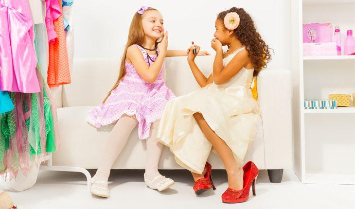 Na foto há duas meninas brincando juntas de se maquiar e vestir roupas como vestidos e saias