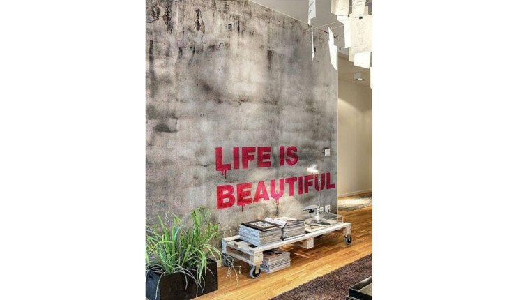 Na foto há uma parede pintada de cimento queimado e manchado com a frase Life is Beautiful em vermelho.