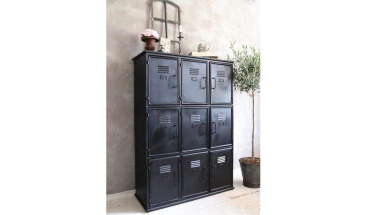 Na foto há um armário de estilo industrial preto com 6 portinhas. Ele está em uma parede pintada de cinza claro, como cor de cimento, e em cima dele há itens decorativos.