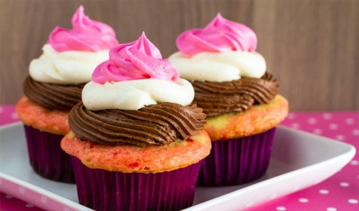 Imagem de um cupcake de massa laranja, cobertura de chocolate e chantily branco e rosa. A forminha é roxa e estão em uma travessa branca.