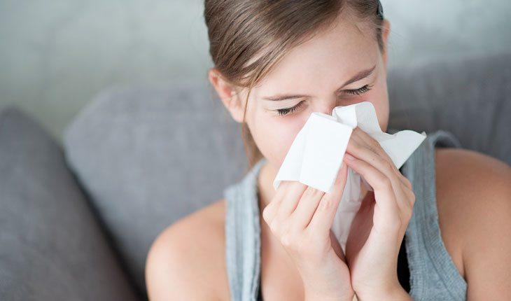 Na imagem há uma mulher espirrando e limpando o nariz com um lenço de papel branco.