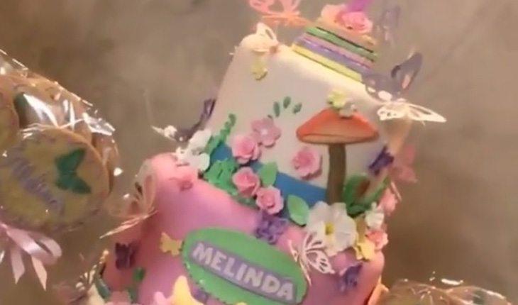 foto do bolo da festa, nas cores rosa e branco, com detalhes, verdes, azuis e lilás. Na primeira camada, está escrito o Melinda e, na segunda, com algumas flores, borboletas e cogumelos