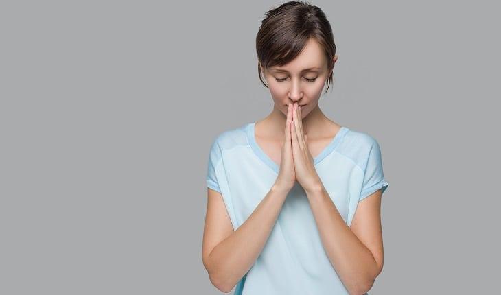 A foto mostra uma mulher com uma camiseta azul clara rezando m pé com as mãos unidas