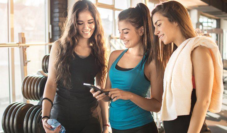 Malhar a dois. Na foto, três amigas estão olhando o celular na mão de uma delas. As três estão sorrindo e em uma academia
