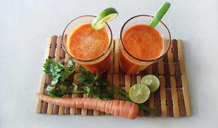 Na foto há dois copos de suco de cenoura na cor laranja com cenoura e limão ao redor.