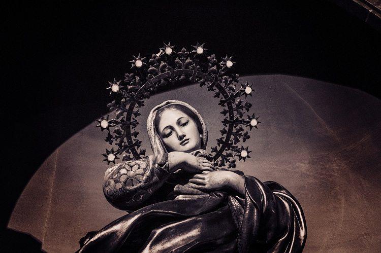 Na imagem, a imagem da virgem maria está de cabeça baixa de olhos fechados com muitas mantas. Virgem maria traz alegria. 