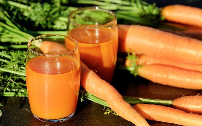 Sucos com colágeno. Na foto, um copo de vitamina de cenoura, com cenouras em volta do copo. Ambos estão em cima de uma bancada de madeira