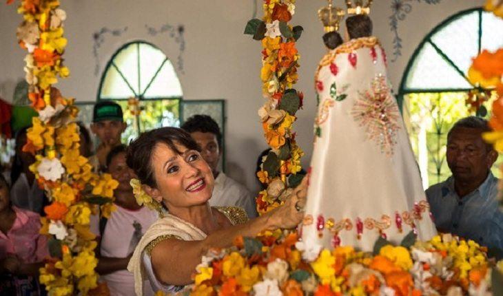 Na imagem, a atriz coloca a mão na Nossa Senhora de Nazaré que está cheia de flores amarelas. A atriz sorri.