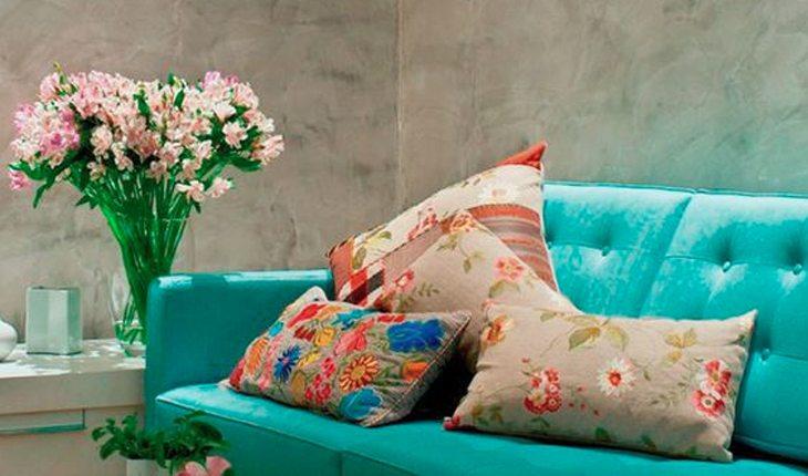 Na foto há um sofá verde-água com almofadas com estampas florais. Ao lado há um vaso de vidro com flores.