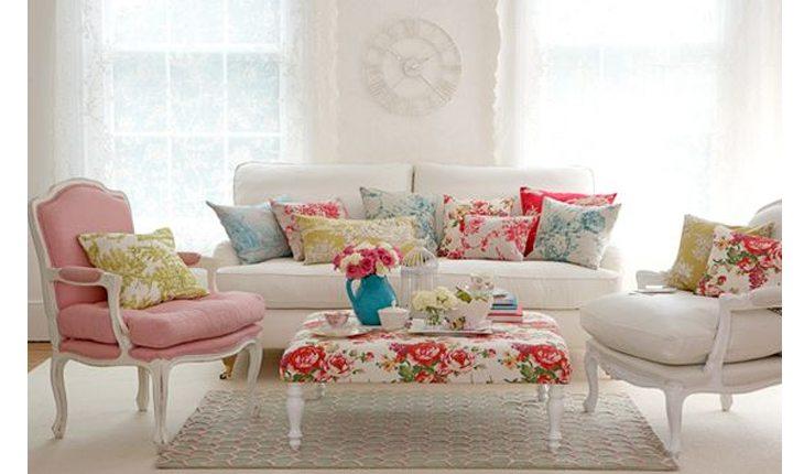 Na foto há um sofá branco com almofadas com estampas florais. De frente há uma banqueta também com estofado floral.
