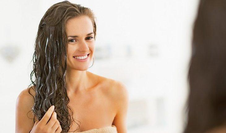 Na foto há uma mulher olhando para o espelho enquanto passa hidratante no cabelo molhado.