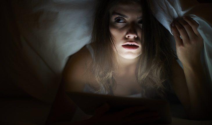 mulher no escuro, iluminada pela luz de um tablet, com um olhar de medo, preocupada