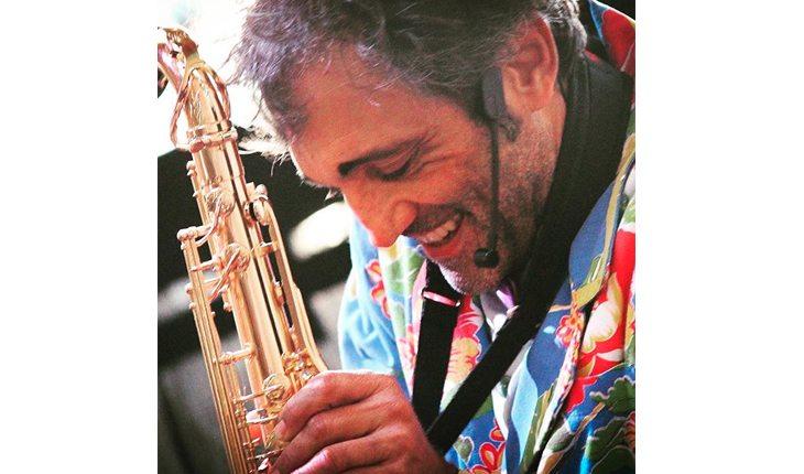 Na foto há o ator Domingos Montagner com uma roupa colorida e um instrumento musical fazendo uma peça