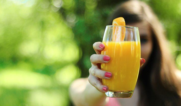 Na foto há uma mulher segurando um copo com suco de laranja