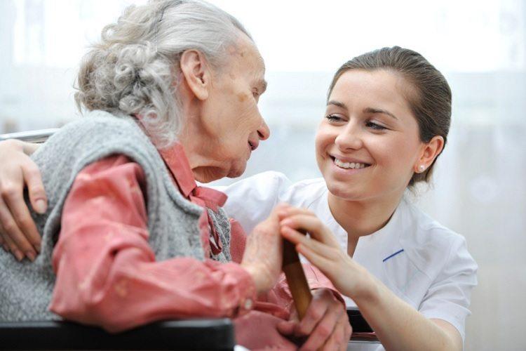 A foto mostra uma mulher sorrindo ajudando uma idosa, demonstrando a importância de um cuidador de idosos saber como agir no dia a dia