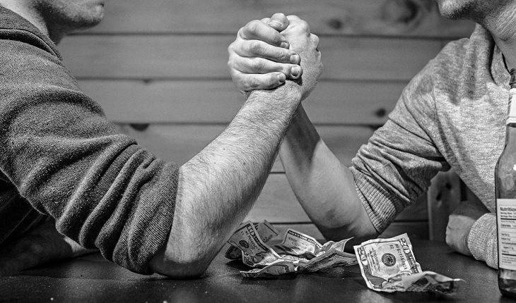 Na imagem, dois homens disputam queda de braço com dinheiro e bebida ao lado. Frases abençoadas.
