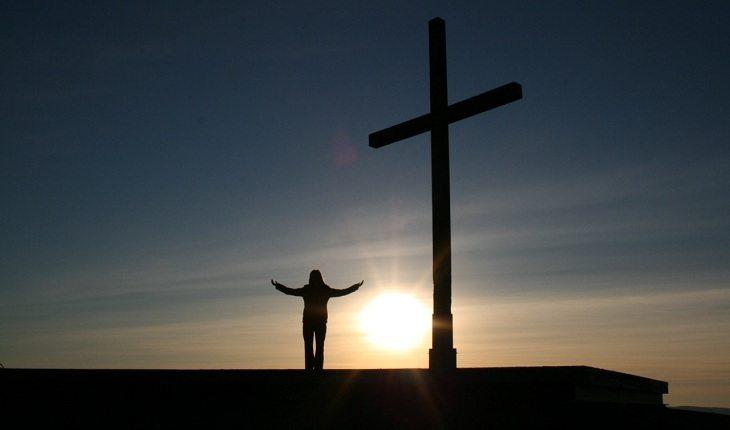 Na imagem, uma pessoa estica os braços em baixo da cruz. Frases abençoadas.