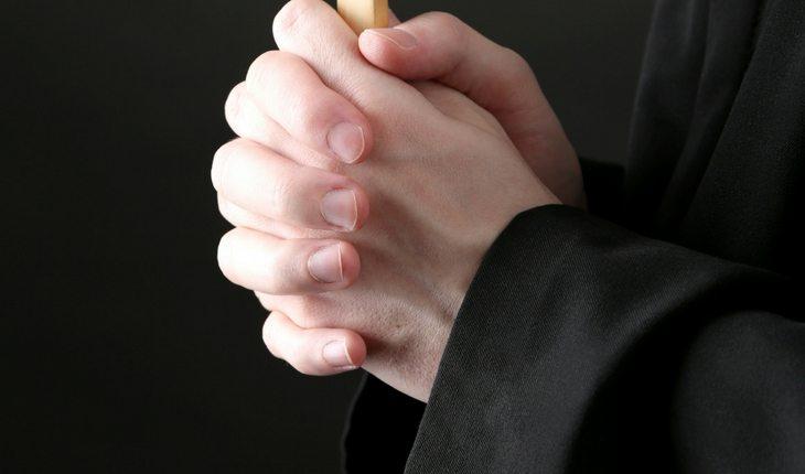 Padre de batina preta rezando com as mãos juntas