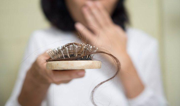Na foto há uma mulher com uma escova de cabelo na mão cheia de cabelos.