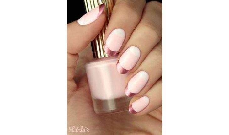Na foto há a mão de uma mulher com as unhas pinadas com um esmalte rosa claro com francesinha em rosa metalizados