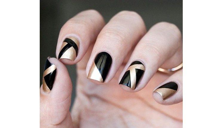 Na foto há a mão de uma mulher com as unhas pinadas com um esmalte preto com detalhes geométricos metalizados