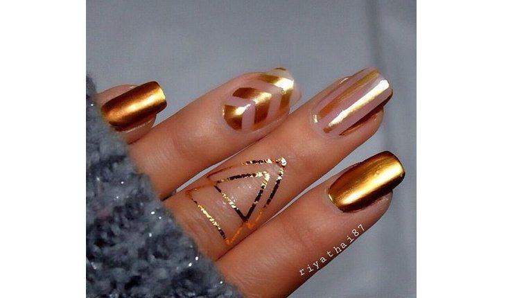 Na foto há a mão de uma mulher com as unhas pinadas com um esmalte metalizado dourado em algumas unhas e com desenhos em rosé gold em outras