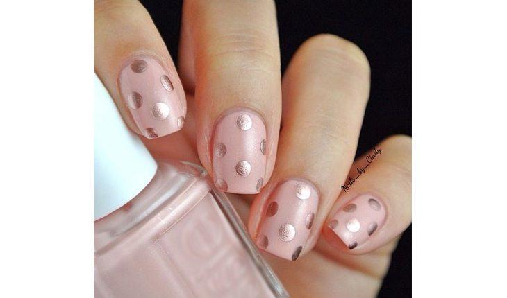 Na foto há a mão de uma mulher com as unhas pinadas com um esmalte rosa-claro com bolinhas metalizadas em rosa