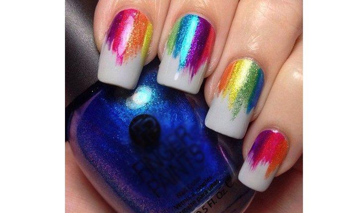Na foto há a mão de uma mulher com as unhas pinadas com um esmalte clarinho e detalhes metalizados nas cores do arco-íris
