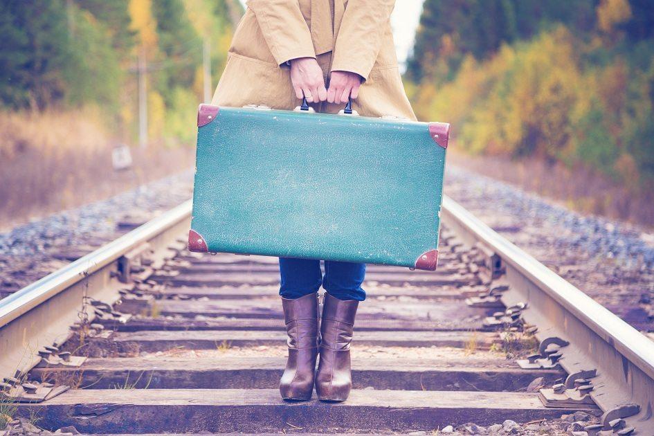 Imagem de uma mulher segurando uma mala azul e antiga com as duas mãos em cima de um trilho de trem.
