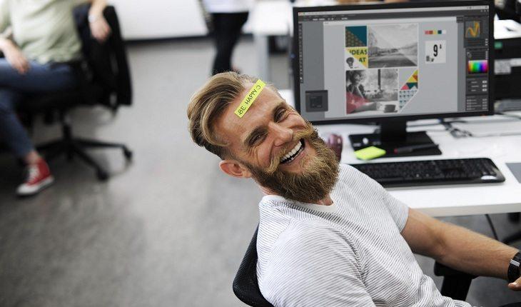 Na imagem, o homem sorri no trabalho com um papel na testa escrito seja feliz. Vida mais feliz.