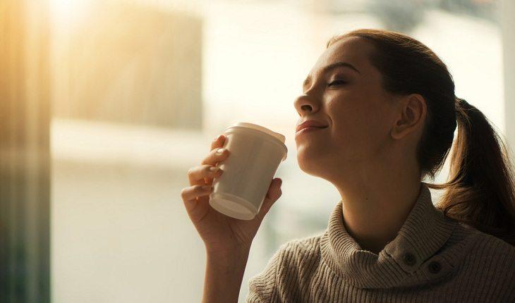 Na imagem, a mulher com olhos fechados sorri para o sol tomando o café de manhã. Vida mais feliz.