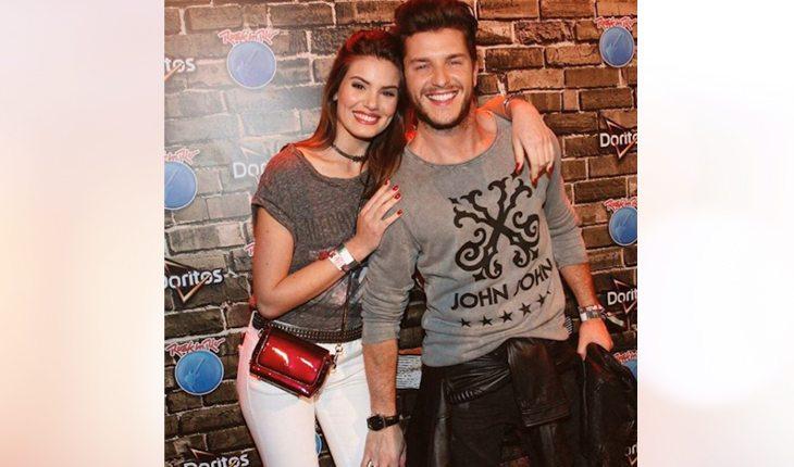 Camila Queiroz e Klebber Toledo em evento a noite. Ela veste uma calça jeans branca com uma blusa cinza enquanto ele usa uma blusa cinza com uma calça preta