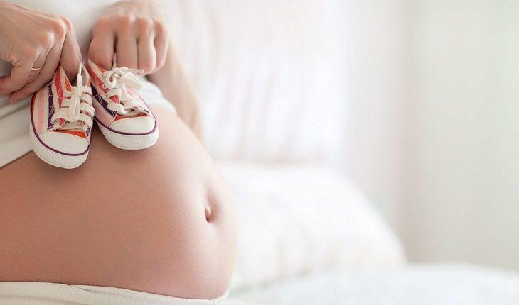 mulher grávida segurando dois sapatinhos de bebê em cima da barriga