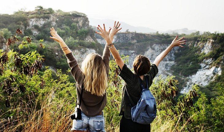 Na imagem, duas amigas erguem os braços para os céus enquanto fazem trilha na natureza. Ensinamentos e reflexões.