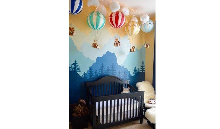 quarto de bebê com a parede decorada com desenhos de montanhas. Acima, há balões decorativos e coloridos pendurados como mobiles.